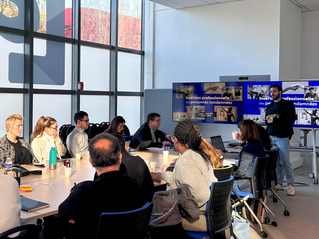 Photo du premier atelier collaboratif post consultation citoyenne Comment améliorer l’insertion professionnelle des personnes condamnées par la justice (formation, emploi, etc.) ? s’est déroulé le 20 mars 2025. Plusieurs personnes asont assises autour d'une table et regardent vers un homme debout devant deux écrans